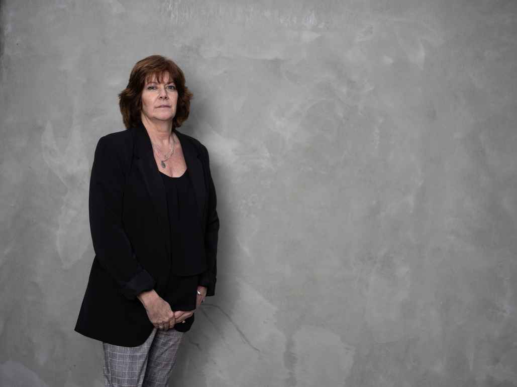 White women wearing a black blazer and grey pants in front of a grey background