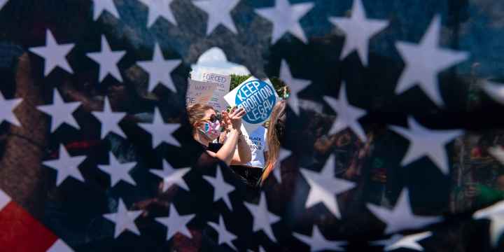 Abortion rights activists are seen through a hole in an American flag as they protest outside the Supreme Court in Washington, Saturday, June 25, 2022.