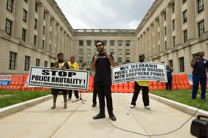 Amol SInha is speaking into a microphone at the State House. Two people are standing behind him holding signs that read "stop police brutality" and "we demand  police review boards with subpoena power"