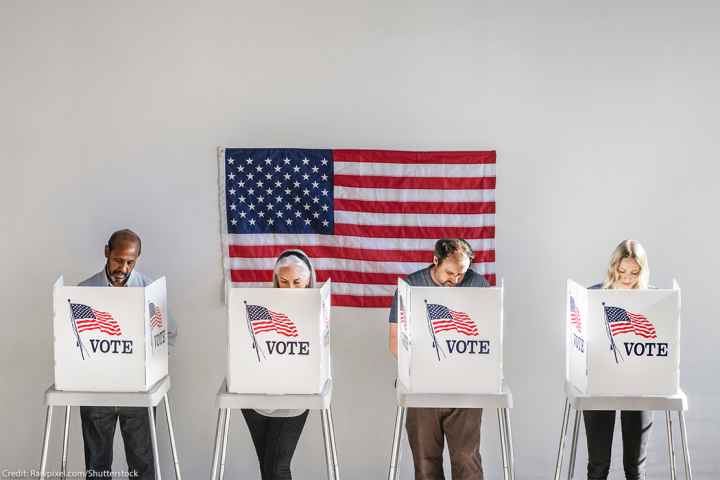 With and American flag in the background, four people in the act of voting, stand behind voter booths as they make their selections.