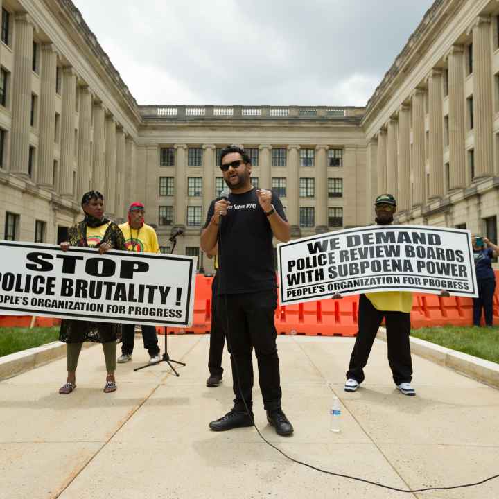 Amol SInha is speaking into a microphone at the State House. Two people are standing behind him holding signs that read "stop police brutality" and "we demand  police review boards with subpoena power"