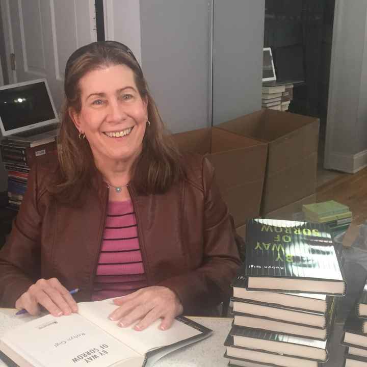 Robyn Gigl smiles for the camera as she sits behind a table of stacked books to her left, and has a book open to sign it.