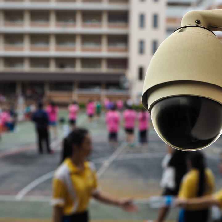 In the foreground, a security camera is situated against a background of students playing during recess